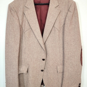 May include: A tan blazer with a subtle plaid pattern, featuring a burgundy interior lining and elbow patches. The blazer has two front pockets, a two-button closure, and buttons on the sleeves. The jacket is hanging on a black hanger.
