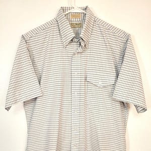 May include: A short-sleeved button-down shirt with a white and black grid pattern. The shirt has a collar, a chest pocket, and is hanging on a white hanger. The brand label "L.L. Bean" is visible.
