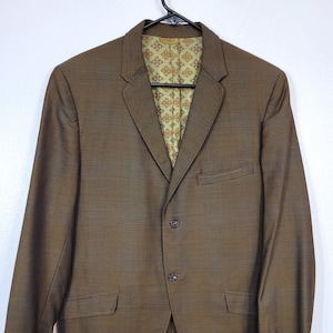 May include: A brown blazer with a textured weave, hanging on a black hanger. The blazer has a three-button closure and two front pockets. The interior lining features a patterned design in shades of green, orange, and brown.