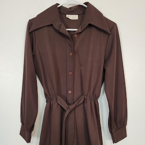 May include: A brown long-sleeved shirt with a collared neckline and button-down front. The shirt features a cinched waist with a matching tie belt. The sleeves have buttoned cuffs. The tag reads "Jill sportswear".