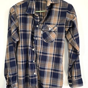 May include: A button-down plaid shirt in navy blue, tan, and light brown. The long-sleeved shirt has a chest pocket and a collar. The shirt is hanging on a green hanger.