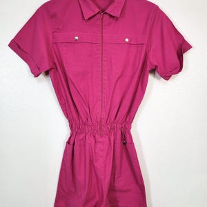 May include: A magenta-colored romper with short sleeves, a collared neckline, and a zip-up front. The romper features two chest pockets with silver button closures and two hip pockets. The waist is cinched with elastic, and the garment hangs on a green hanger.