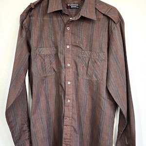 May include: Brown button-up shirt with vertical striped pattern. Features two chest pockets with decorative studs and epaulettes on the shoulders. The shirt has long sleeves and a classic collar. The label reads "AMERICAN VOGUE".