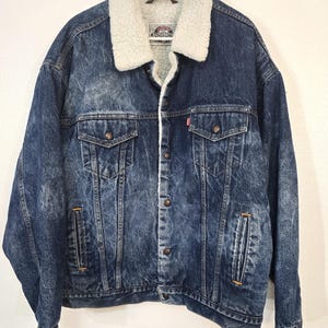 May include: A dark blue acid-wash denim jacket featuring a thick white sherpa fleece collar and lining. The jacket includes two chest pockets with button-down flaps, two vertical side pockets, and a front button closure with metallic hardware.