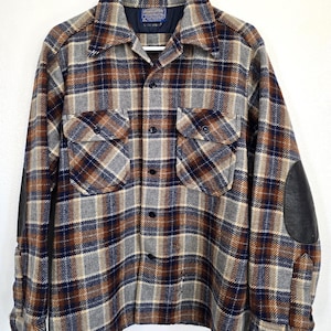 May include: A long-sleeve button-up shirt featuring a plaid pattern in shades of brown, navy blue, and gray. The shirt includes two chest pockets with button flaps, a pointed collar, and black leather-like elbow patches on each sleeve.