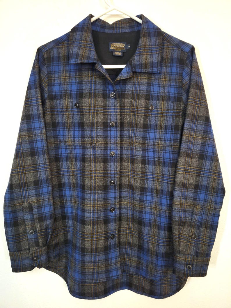 K&ouml;nnte beinhalten: Ein blaues, schwarzes und graues kariertes Button-Down-Hemd. Das Hemd hat lange &Auml;rmel, zwei Brusttaschen und einen Kragen. Das Pendleton-Markenetikett ist auf der Innenseite des Kragens sichtbar. Das Hemd h&auml;ngt an einem wei&szlig;en Kleiderb&uuml;gel.