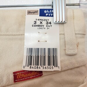 May include: Close-up of off-white Wrangler Cowboy Cut jeans, size 3 x 34, hanging on a hanger. A tag displays the size and brand information. The Wrangler logo is visible on a red patch.