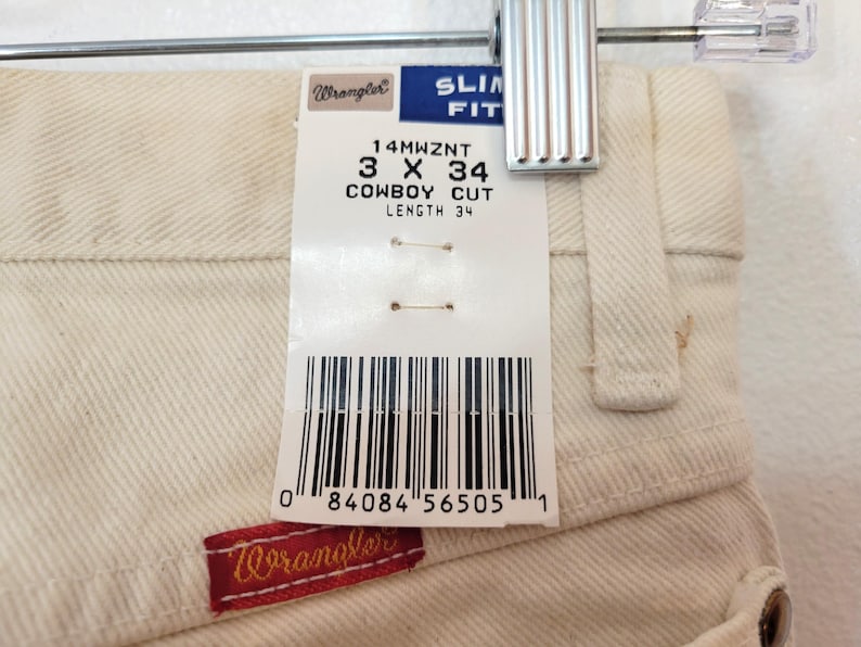 May include: Close-up of off-white Wrangler Cowboy Cut jeans, size 3 x 34, hanging on a hanger. A tag displays the size and brand information. The Wrangler logo is visible on a red patch.