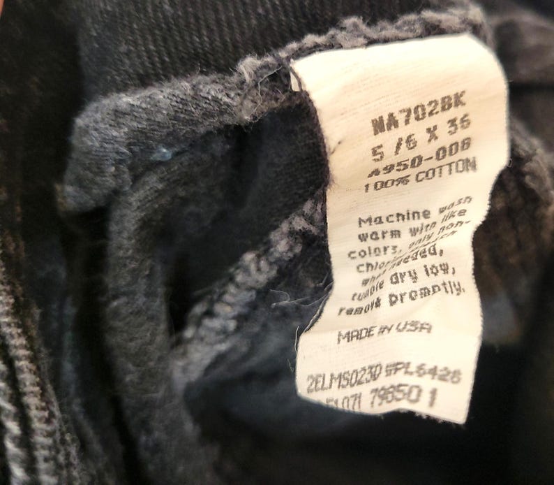Puede incluir: Primer plano de una prenda de mezclilla negra con una etiqueta blanca. La etiqueta muestra texto que incluye "NA702BK", "5/6 X 36" y "100% COTTON". La etiqueta tambi&eacute;n incluye instrucciones de lavado y "MADE IN USA".