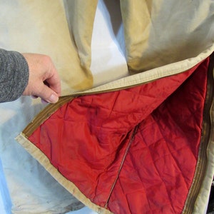 May include: A pair of tan work pants with a red quilted lining. The pants have a zipper on the side and a brown zipper pull.