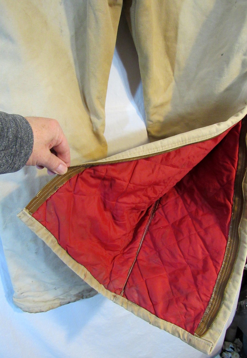 May include: A pair of tan work pants with a red quilted lining. The pants have a zipper on the side and a brown zipper pull.