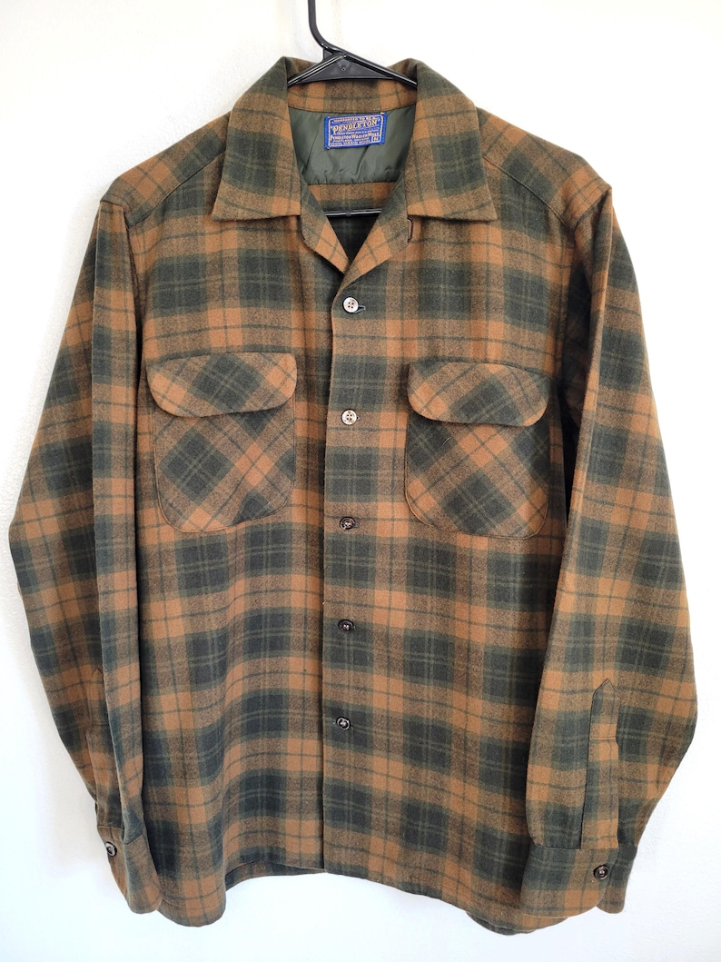 Pode incluir: Uma camisa de manga comprida com bot&otilde;es, com um padr&atilde;o xadrez castanho e verde. A camisa tem dois bolsos no peito e uma gola. Est&aacute; pendurada num cabide preto sobre um fundo branco.