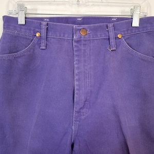 May include: Close-up of a pair of purple denim jeans. The jeans have a button and zipper closure, front and back pockets, and belt loops. The jeans are hanging on a white hanger.
