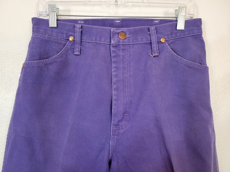 May include: Close-up of a pair of purple denim jeans. The jeans have a button and zipper closure, front and back pockets, and belt loops. The jeans are hanging on a white hanger.