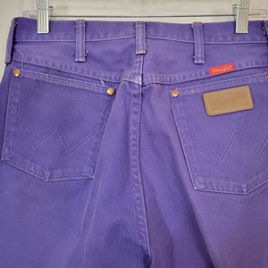 May include: Close-up of the back of purple Wrangler jeans. The jeans have two back pockets with the signature Wrangler stitching, copper rivets, and a brown leather patch. The Wrangler logo is on a red tag.