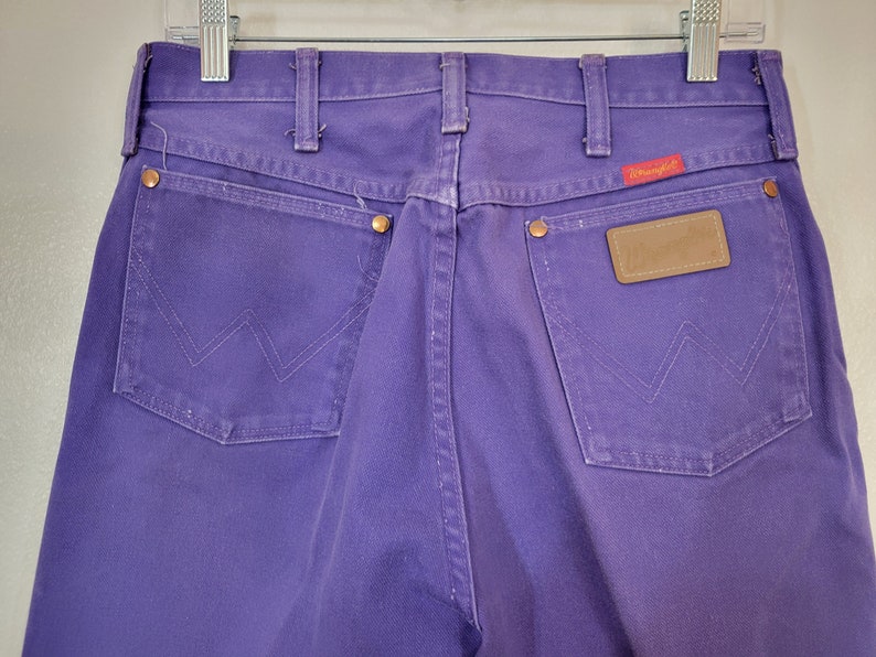 May include: Close-up of the back of purple Wrangler jeans. The jeans have two back pockets with the signature Wrangler stitching, copper rivets, and a brown leather patch. The Wrangler logo is on a red tag.