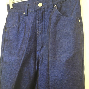 May include: Close-up of a pair of dark blue denim pants. The pants have a button and zipper closure, front and back pockets, and belt loops. The denim has a subtle sheen.