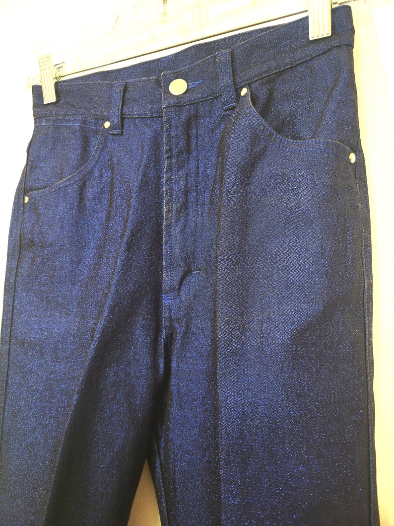 May include: Close-up of a pair of dark blue denim pants. The pants have a button and zipper closure, front and back pockets, and belt loops. The denim has a subtle sheen.
