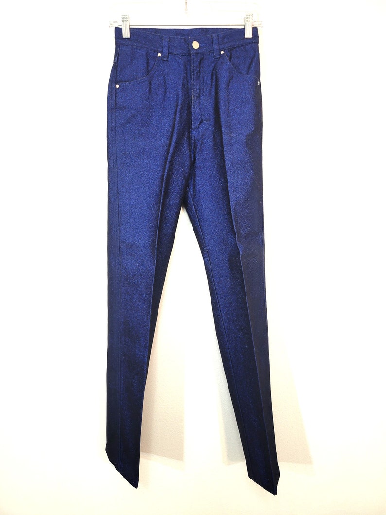 May include: A pair of dark blue denim pants hanging on a white hanger. The pants have a classic five-pocket design with a button and zipper closure. The denim has a subtle sheen, and the pants appear to be new.