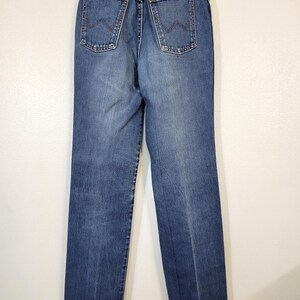 May include: Back view of blue denim jeans with a classic straight leg cut. The jeans feature two back pockets with a distinctive stitched design, a brown leather patch, and a metal button closure. The jeans are hanging on a white hanger.