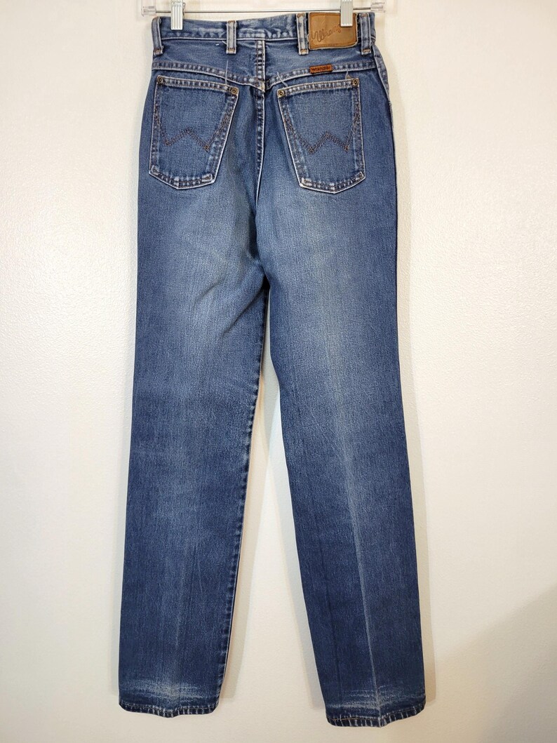 May include: Back view of blue denim jeans with a classic straight leg cut. The jeans feature two back pockets with a distinctive stitched design, a brown leather patch, and a metal button closure. The jeans are hanging on a white hanger.