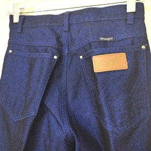 May include: Close-up of dark blue Wrangler jeans. The jeans have a classic five-pocket design with a leather patch and the Wrangler logo. The back pockets feature the signature Wrangler stitching.