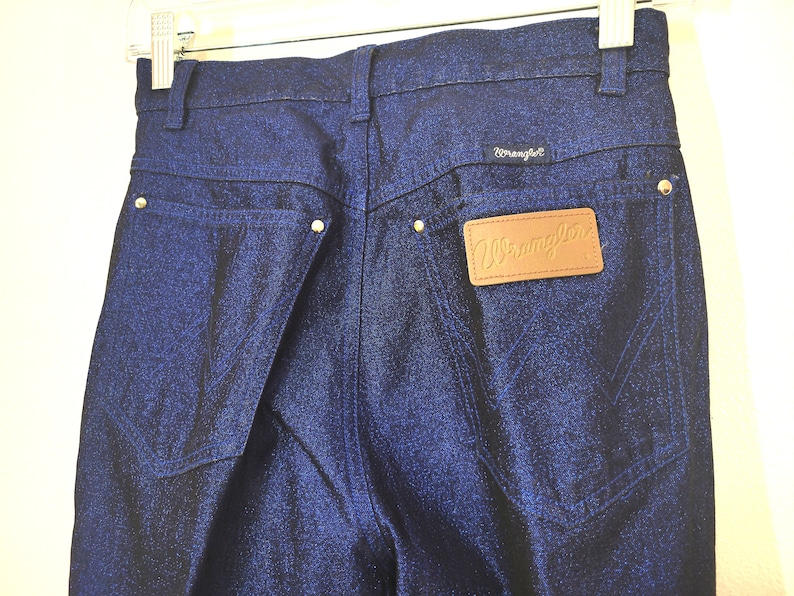 May include: Close-up of dark blue Wrangler jeans. The jeans have a classic five-pocket design with a leather patch and the Wrangler logo. The back pockets feature the signature Wrangler stitching.