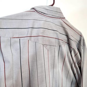 May include: A light gray button-up shirt with vertical stripes in black and burgundy. The shirt is hanging on a brown hanger. The back of the shirt is visible, showing the yoke and collar.