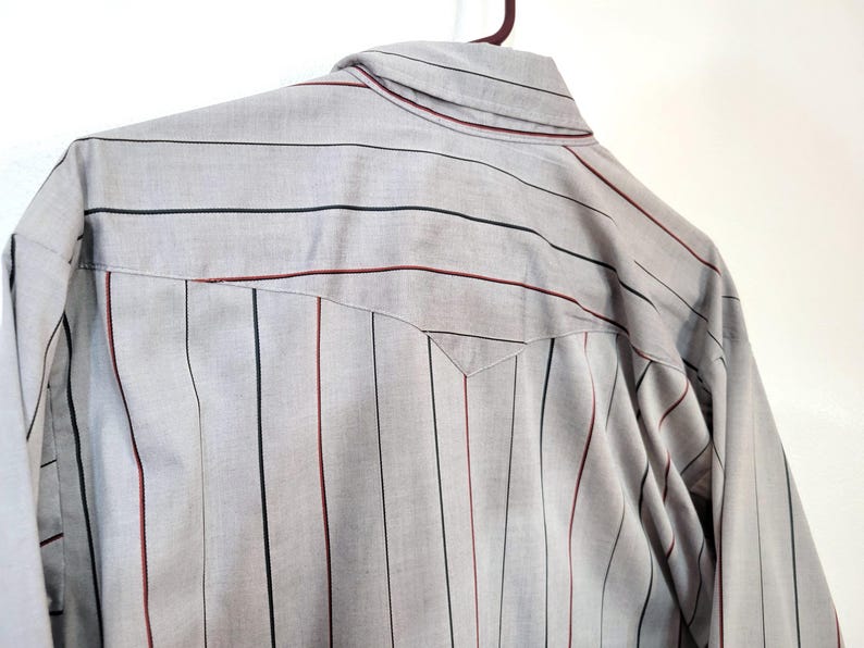 May include: A light gray button-up shirt with vertical stripes in black and burgundy. The shirt is hanging on a brown hanger. The back of the shirt is visible, showing the yoke and collar.