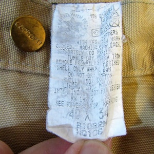 May include: A tan Carhartt workwear label with white text. The label includes care instructions, size information, and the text "Crafted with Pride".