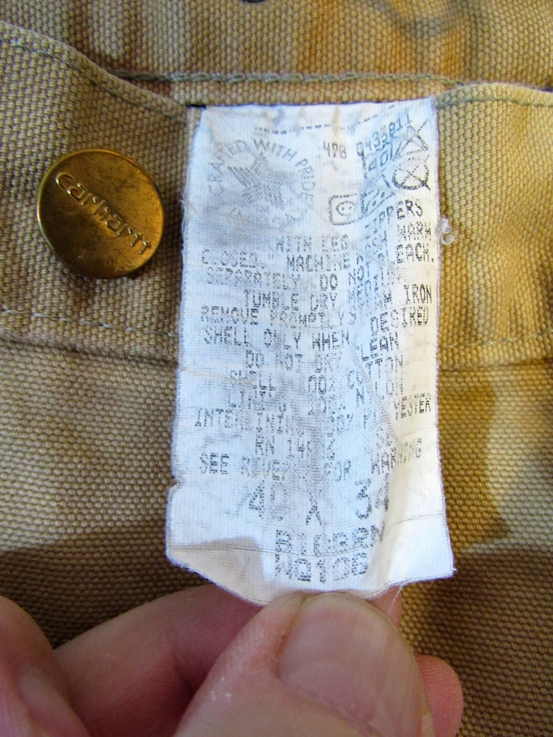 May include: A tan Carhartt workwear label with white text. The label includes care instructions, size information, and the text "Crafted with Pride".