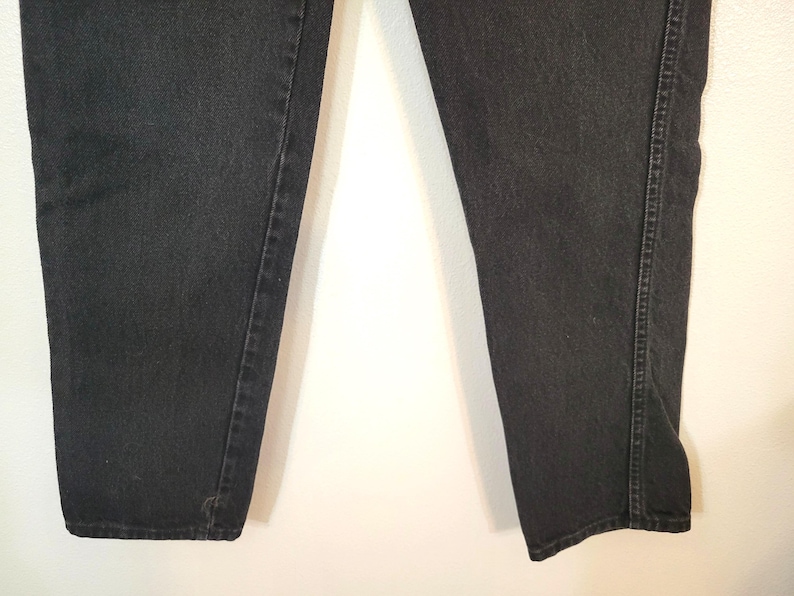 Puede incluir: Primer plano de unos vaqueros de mezclilla negros. Los vaqueros tienen un dise&ntilde;o cl&aacute;sico de cinco bolsillos, con un corte de pierna recta. El denim es de color negro liso y las costuras son visibles a lo largo de las costuras.