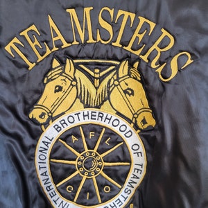 May include: Black satin jacket with gold embroidered "TEAMSTERS" lettering and a logo featuring two horses and a wagon wheel. The logo also includes the text "INTERNATIONAL BROTHERHOOD OF TEAMSTERS AFL CIO".