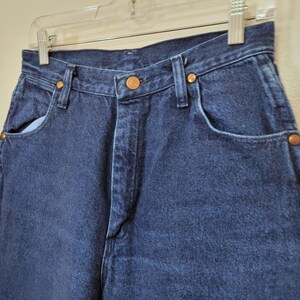 May include: Close-up of dark blue denim jeans. The jeans have a high waist, classic five-pocket design, and copper-colored rivets. The jeans are hanging on a clear plastic hanger against a neutral background. The denim appears to be a medium weight.