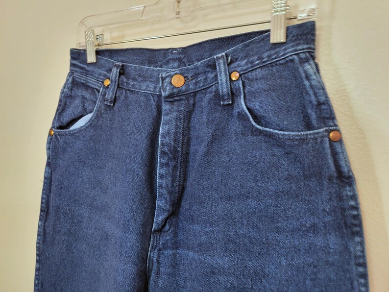 May include: Close-up of dark blue denim jeans. The jeans have a high waist, classic five-pocket design, and copper-colored rivets. The jeans are hanging on a clear plastic hanger against a neutral background. The denim appears to be a medium weight.