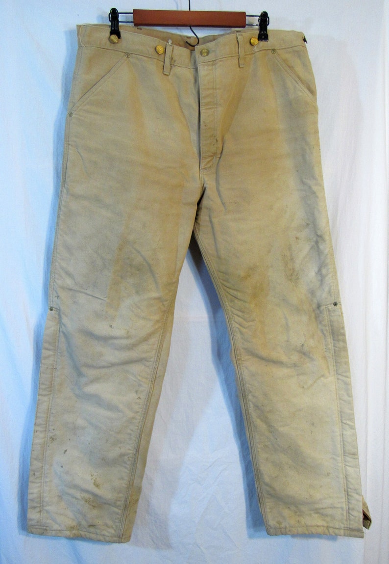 May include: A pair of tan work pants with a worn and faded appearance. The pants have multiple pockets and a button fly.