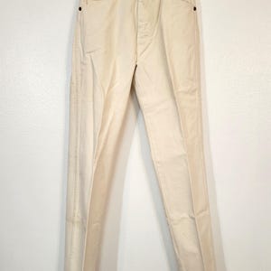 May include: Cream-colored denim pants hanging on a clear hanger. The pants have a button and zipper closure, front and back pockets, and belt loops. The pants are a light, neutral color.