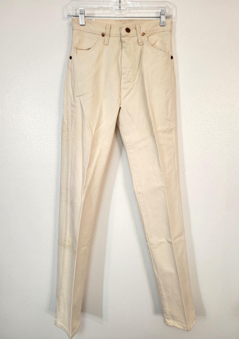 May include: Cream-colored denim pants hanging on a clear hanger. The pants have a button and zipper closure, front and back pockets, and belt loops. The pants are a light, neutral color.