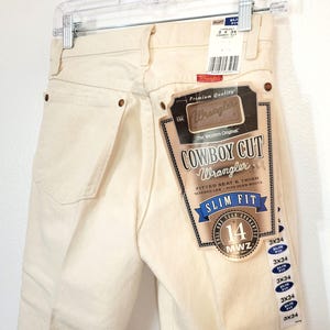 May include: Cream-colored Wrangler Cowboy Cut Slim Fit jeans, size 14, with a fitted seat and thigh, tapered leg, and designed to fit over boots. The jeans have a branded tag and are hanging on a hanger.
