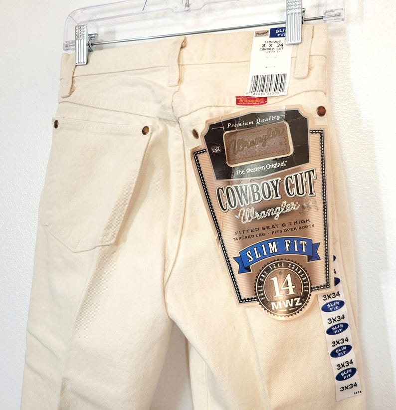 May include: Cream-colored Wrangler Cowboy Cut Slim Fit jeans, size 14, with a fitted seat and thigh, tapered leg, and designed to fit over boots. The jeans have a branded tag and are hanging on a hanger.