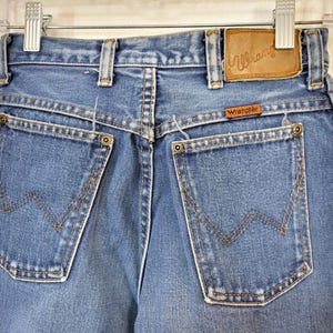 May include: Close-up of the back of blue denim Wrangler jeans. The jeans have two back pockets with the Wrangler logo stitched on them. A brown leather patch with the Wrangler logo is visible. The jeans are hanging on a metal hanger.