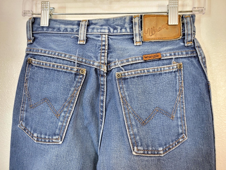 May include: Close-up of the back of blue denim Wrangler jeans. The jeans have two back pockets with the Wrangler logo stitched on them. A brown leather patch with the Wrangler logo is visible. The jeans are hanging on a metal hanger.