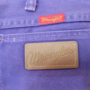 May include: Close-up of purple denim jeans featuring a back pocket with a brown leather Wrangler label. The Wrangler logo is also visible on a red tag. Copper rivets and white stitching detail the jeans.