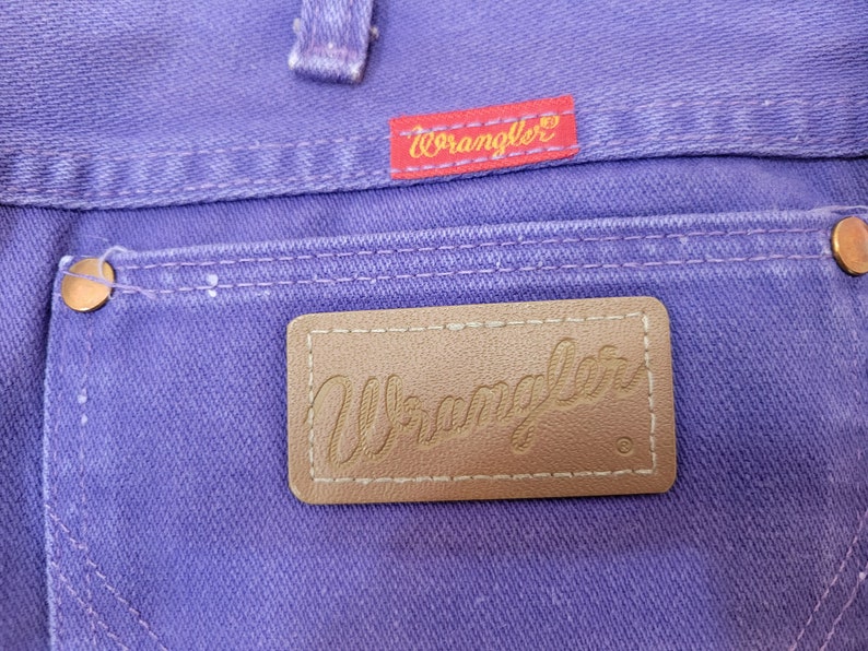 May include: Close-up of purple denim jeans featuring a back pocket with a brown leather Wrangler label. The Wrangler logo is also visible on a red tag. Copper rivets and white stitching detail the jeans.