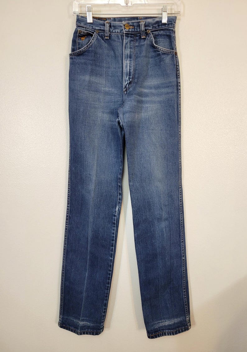 May include: Vintage blue denim jeans with a high waist and straight leg. The jeans have a classic five-pocket design, a button and zipper closure, and belt loops. The denim has a faded appearance.