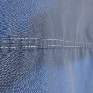 May include: Close-up of blue fabric with two parallel rows of light green stitching. The fabric has a textured appearance, and the stitching is evenly spaced, creating a clean, tailored look. The image highlights the details of the textile.