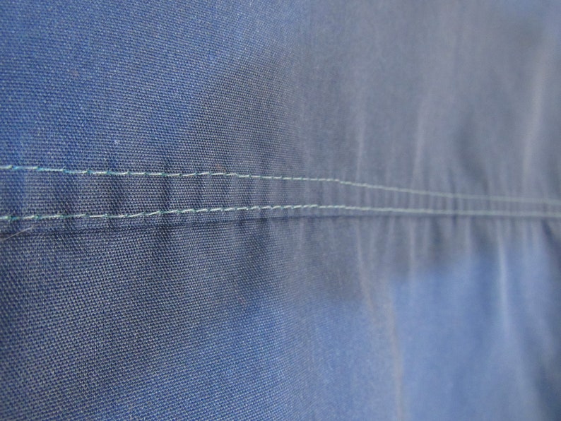 May include: Close-up of blue fabric with two parallel rows of light green stitching. The fabric has a textured appearance, and the stitching is evenly spaced, creating a clean, tailored look. The image highlights the details of the textile.