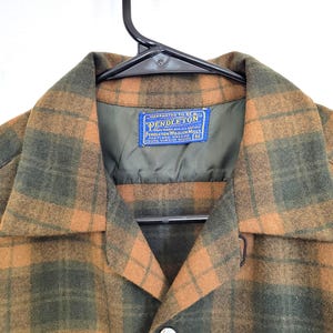 Pode incluir: Uma camisa de tamanho m&eacute;dio, com bot&otilde;es, em um padr&atilde;o xadrez verde e laranja. A camisa est&aacute; pendurada em um cabide preto. A etiqueta diz "Pendleton Woolen Mills".