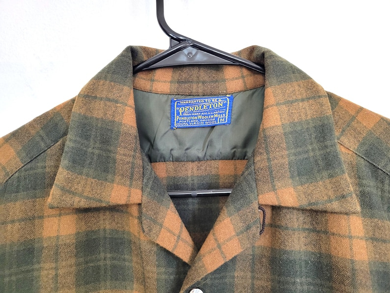Pode incluir: Uma camisa de tamanho m&eacute;dio, com bot&otilde;es, em um padr&atilde;o xadrez verde e laranja. A camisa est&aacute; pendurada em um cabide preto. A etiqueta diz "Pendleton Woolen Mills".