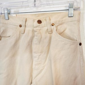 May include: Close-up of cream-colored denim jeans hanging on a clear hanger. The jeans have a classic five-pocket design with copper-colored rivets and a button closure. The fabric appears slightly worn.